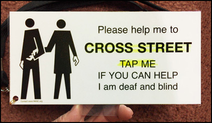The card has a drawing of one person guiding another, and says: 'Please help me to -- CROSS STREET -- TAP ME -- IF YOU CAN HELP -- I am deaf and blind.' The words 'Cross street, tap me' are highlighted in yellow.
The card has small holes at the top right and bottom left; a neck cord attached to the bottom left corner. The card is held by a see-through tab at the bottom.